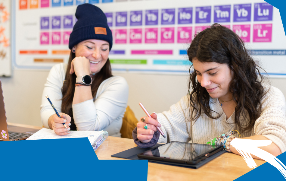 Two students working together at a classroom table, with one taking notes and the other using a stylus on a tablet, while a periodic table is displayed on the wall behind them.